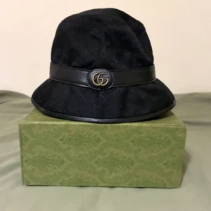 Svart bucket hat från Gucci - Snygg svart bucket hat från Gucci med GG-logga i guld framtill. Hatten har ett brett läderband runtom och är tillverkad i ett tyg med diskret GG-mönster. Perfekt accessoar för att lyfta din outfit med lyxig känsla.