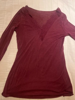 Vinröd långärmad blus med spets (S) - Säljer en vinröd långärmad blus från Intimissimi med v-ringad hals och dekorativ spets både vid halsringningen och ärmsluten. Tunn och mjuk i materialet, perfekt för dig som gillar romantiska detaljer och snygg passform.
