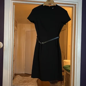 Svart kort klänning med bälte - Snygg svart kort klänning med rund hals och korta ärmar. Klänningen har ett coolt bälte med metallspänne i midjan som ger en edgy vibe. Dragkedja bak för enkel påklädning. Passformen är normal och materialet känns som polyester.