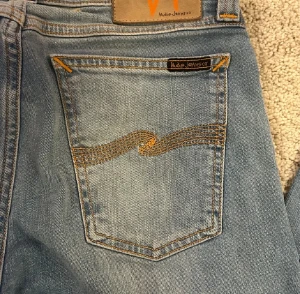 Blå jeans från Nudie Jeans - Säljer ett par klassiska blå jeans från Nudie Jeans med snygga orangea sömmar och broderad våg på bakfickorna. Jeansen har rak passform och normal midja, tillverkade i mjukt bomullsdenim. Perfekt för dig som gillar stilrena och tidlösa jeans.