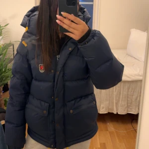 Blå dunjacka från Fjällräven - Säljer en blå dunjacka från Fjällräven med huva och gula snören. Jag köpte denna jacka förra året, den är i storlek XS. Säljer på grund av att jag ska köpa en ny i en annan färg.  Perfekt för kalla dagar och har en riktigt mysig, puffig känsla. Inga defekter, men smink vid luvan, skriv för fler bilder.