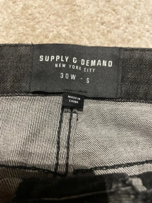 Svarta jeans från Supply & Demand, 30W - Svarta jeans från Supply & Demand med slitna detaljer och färgstänk på framsidan. Modellen har fem fickor och smal passform. Jeansen är tillverkade i jeansmaterial och har en cool, urban vibe med distressed look.