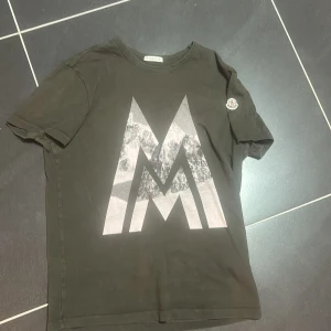 Mörkgrön Moncler t-shirt  - Cool mörkgrön t-shirt från Moncler med stor grafisk M-print på framsidan och Moncler-logga på ärmen. Rund hals och klassisk passform. Materialet är mjuk bomull som känns skönt mot huden. Passar S 