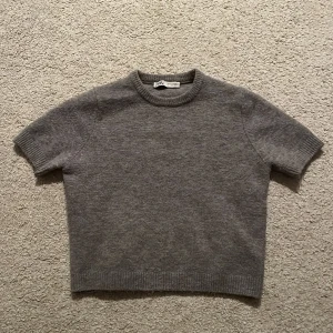 Grå stickad tröja från Zara - Säljer en enkel och stilren grå stickad tröja från Zara i storlek M. Tröjan har korta ärmar, rund hals och ribbade muddar vid hals, ärmslut och nederkant. Perfekt för dig som gillar minimalistisk och clean look.