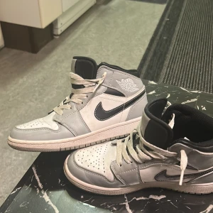 Nike Air Jordan 1 Mid grå/vit - Nike Air Jordan 1 Mid sneakers i grått och vitt med svarta detaljer och klassisk Swoosh på sidan. Skorna har snörning, perforerad tå och högt skaft. Materialet är skinn och syntet, med en platt sula och rund tå. Perfekt för dig som gillar streetstyle och ikoniska sneakers.