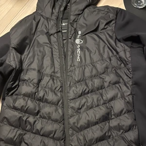 Svart pufferjacka från Sail Racing - Svart pufferjacka från Sail Racing med huva och dragkedja. Jackan har quiltad design, vind- och vattenavvisande material med Primaloft-foder. Snygg logga på bröstet och huvan. Perfekt för kyliga dagar och har en sportig vibe. Den är äkta