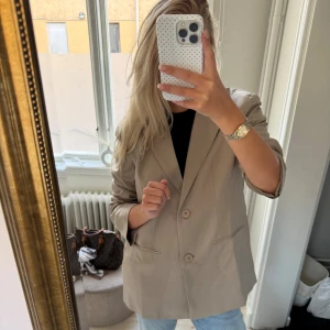 Beige kavaj med knappar - Snygg beige kavaj med två knappar framtill och klassisk krage. Modellen är rak och lite oversized, perfekt att styla med jeans eller dressade byxor. Kavajen har fickor på sidorna och är tillverkad i ett slätt material som ger en clean look.