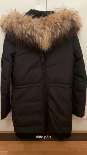 Svart dunjacka med äkta päls från Hollies - Snygg svart dunjacka från Hollies med stor fluffig äkta päls runt huvan. Jackan har guldiga detaljer, dragkedja och knappar framtill samt en bröstficka med dragkedja. Perfekt för kalla vinterdagar och riktigt stilren look. Priset kan även diskuteras!