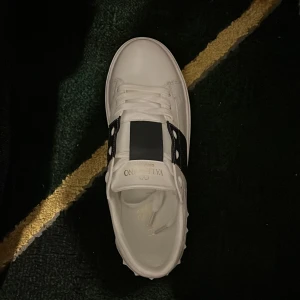 Valentino Open sneakers vit/svart - Säljer ett par Valentino Open sneakers i vitt skinn med svart bred rem över vristen och ikoniska nitar på hälen. Klassisk rund tå, platt sula och snörning. Perfekta för dig som vill ha en clean men lyxig look.