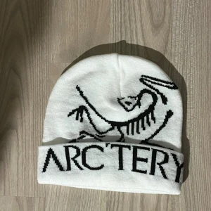 Vit mössa från Arcteryx - Snygg vit mössa från Arcteryx med svart logga och stor text framtill. Klassisk modell i mjukt material som håller dig varm och passar perfekt till kalla dagar. Enkel att matcha med olika outfits och riktigt clean look.