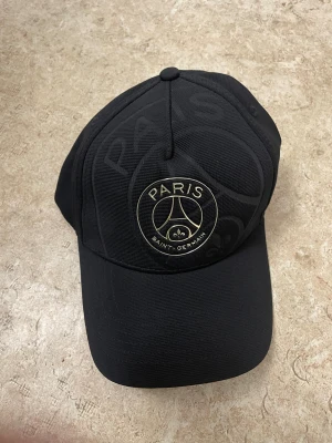 Svart PSG keps med guldtryck - Snygg svart keps från Paris Saint-Germain med guldigt tryck på framsidan och texten 'ICI C'EST PARIS' broderad baktill. Justerbar rem och böjd skärm. Materialet är slitstark polyester och insidan har PSG-logga tryckt längs kanten.