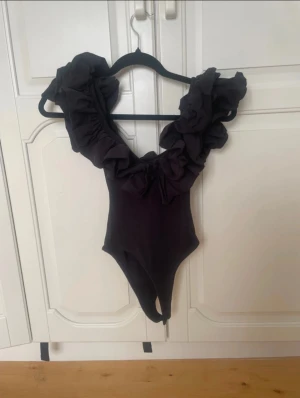 Svart body med volangdetaljer - Supersnygg svart body med djup v-ringning och stora volanger längs axlar och urringning. Bodyn har öppen rygg och en stilren, elegant look som verkligen passar till fest eller vardag. 