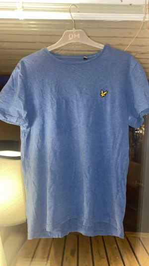 Blå t-shirt från Lyle & Scott, M - Snygg blå t-shirt från Lyle & Scott i storlek M, passar på S. Klassisk rund hals och korta ärmar. På bröstet sitter den ikoniska gula fågel-loggan. Tillverkad i mjuk bomull som känns skön mot huden. Perfekt för dig som gillar stilrena och enkla plagg.