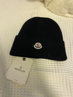 Svart Moncler mössa i ull - Svart ribbstickad mössa från Moncler med klassisk logotyp framtill. Snygg och enkel design som passar till allt, tillverkad i mjuk ull för extra värme. Perfekt för kalla dagar och en stilren look.
