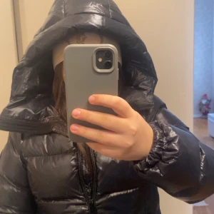 Svart pufferjacka från Everest - Säljer en svart, glansig pufferjacka från Everest med stor huva och dragkedja framtill. Jackan har ett snyggt badge på ärmen och är riktigt fluffig och varm. Perfekt för kalla dagar när du vill ha en cool och sportig look.