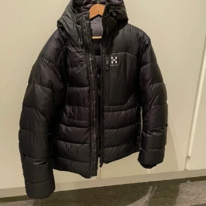 Svart dunjacka från Haglöfs - Svart dunjacka från Haglöfs med huva och markerad logga på bröstet. Jackan har quiltad design, dragkedja framtill och två sidofickor med dragkedja. Perfekt för kalla dagar och har en schysst, lite puffig look som är både snygg och praktisk.
