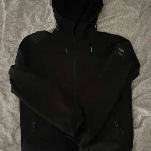 Svart vindjacka från Jack & Jones - Svart vindjacka från Jack & Jones med huva och dragsko, hel dragkedja framtill och två fickor med dragkedja. Snygg patch på ärmen med koordinater. Perfekt för dig som gillar stilren och enkel design.