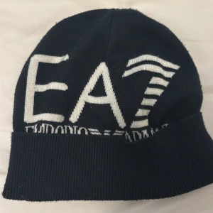 EA7 Emporio Armani mörkblå mössa - Snygg mörkblå mössa från EA7 Emporio Armani med stor vit logga framtill. Mössan är stickad i mjuk akryl och har en bred ribbad kant för extra komfort. Perfekt för dig som gillar stilrena accessoarer med tydlig branding.