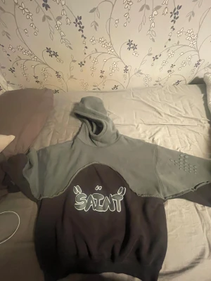 Grå och svart hoodie med SAINT-tryck - Säljer en unik hoodie från Saint i grått och svart med coolt grafiskt SAINT-tryck på bröstet. Hoodien har huva, lång ärm med detaljerade prickar på ena ärmen och råa sömmar som ger en edgy vibe. Perfekt för dig som gillar streetwear och vill sticka ut.