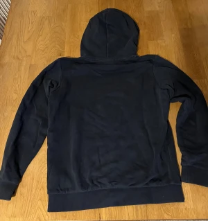 Mörkblå hoodie från Peak Performance - Snygg mörkblå hoodie från Peak Performance med vit broderad logga på bröstet. Tröjan har huva, känguruficka och ribbade muddar vid ärmslut och nederkant. Perfekt för chill eller när du vill ha en enkel stil.
