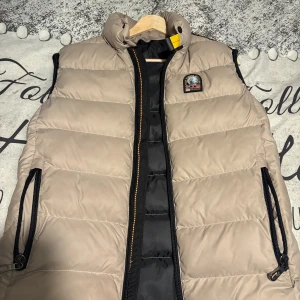 Beige dunväst från Parajumpers - Snygg beige dunväst från Parajumpers med svart dragkedja och coolt patch på bröstet. Västen har hög krage med gul detalj, två fickor med dragkedja och quiltad design. Perfekt för att addera lite edge till din outfit.