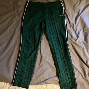 Gröna mönstrade träningsbyxor Lacoste - Snygga gröna träningsbyxor från Lacoste med geometriskt mönster och vita ränder längs sidorna. Byxorna har elastisk midja och den klassiska krokodilloggan på benet. Regular fit och sportig vibe, perfekta för chill eller träning.