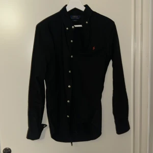 Svart skjorta Polo Ralph Lauren - Svart skjorta från Polo Ralph Lauren i slim fit med klassisk krage och vita knappar. Broderad röd logga på bröstet. Tillverkad i mjuk bomull, långärmad och stilren design som funkar till många olika looks.