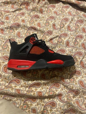 Svarta och röda Nike Air Jordan 4 - Nike Air Jordan 4 sneakers i svart och rött med klassisk Jumpman-logga på hälen och sulan. Skorna har snörning, meshpaneler på sidorna och detaljer i plast. Sulan är platt med synlig luftkudde. Materialet är mestadels skinn och syntet.
