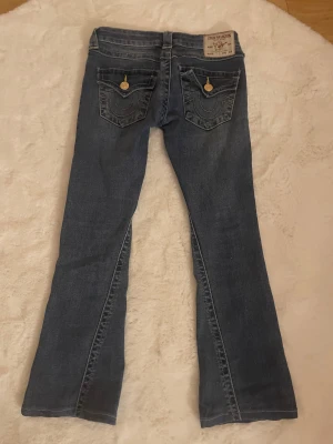 True Religion Joey Low Rise Flare Jeans - Snygga blå jeans från True Religion, modell Joey Low Rise Flare. Byxorna har låg midja, utsvängda ben och klassiska fickor med gul kontrastsöm och dekorativa knappar. Tillverkade i jeansmaterial med coola detaljer och ikonisk logga bak. Passar 152 har sytt upp dem