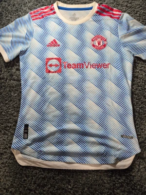 Manchester United bortatröja Adidas M - Manchester Uniteds bortatröja från Adidas i storlek M. Tröjan har ett diagonalrandigt mönster i vitt och blått, röda detaljer och loggor, samt klassiska röda Adidas-ränder på axlarna. Rund hals och korta ärmar. Materialet är lätt och andas, perfekt för match eller träning.