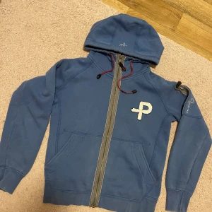 Blå hoodie från Pelle P med dragkedja - Säljer en blå hoodie från Pelle P med hel dragkedja, huva och röda snören. Tröjan har broderad logga på bröstet och ärmen, samt praktiska fickor framtill. Perfekt för dig som gillar sportig och avslappnad stil.