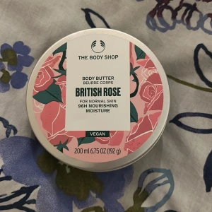 The Body Shop British Rose Body Butter - En vegansk body butter med doft av British Rose från The Body Shop. Kommer i en rund burk med rosa och grönt blommigt lock, innehåller 200 ml (192 g) krämig, vit fuktgivande formula som ger näring i upp till 96 timmar. Perfekt för normal hud och ger en fräsch rosendoft.
