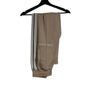 Palm Angels beige mjukisbyxor - Beige mjukisbyxor från Palm Angels med vita och svarta ränder längs sidorna. Byxorna har elastiska muddar vid bensluten och Palm Angels-logga tryckt på ena benet. Snyggt sportig look och skön passform.