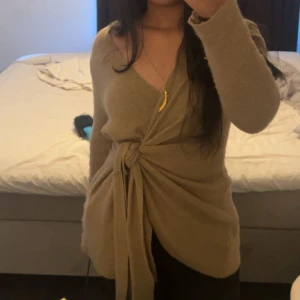 Beige omlott kofta med knyt - Säljer en beige omlott kofta med snygg knyt framtill och v-ringad hals. Koftan har långärmade ärmar och är gjord i ett mjukt stickat material som känns skönt mot huden. Perfekt för dig som gillar chill och stilren look. Perfekt nu till vintern, där den både värmer dig och får dig att se lite bättre ut under vardagen! 🤭