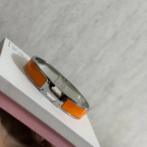  orange H-armband i metall - Snyggt och stilrent armband med klassisk H-detalj framtill. Armbandet är i blankt silverfärgat metall med orange emaljpaneler runt om. Perfekt statement piece för dig som gillar färg och exklusiv design.