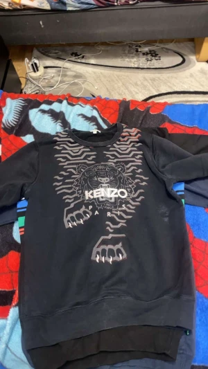 Svart Kenzo sweatshirt med tigerprint - Svart sweatshirt från Kenzo med grymt tigertryck och Paris-text på bröstet. Tröjan har långärmade och en klassisk rund hals. Snyggt grafiskt mönster i grått och silver som gör den riktigt unik. Perfekt för dig som gillar streetwear och vill sticka ut.