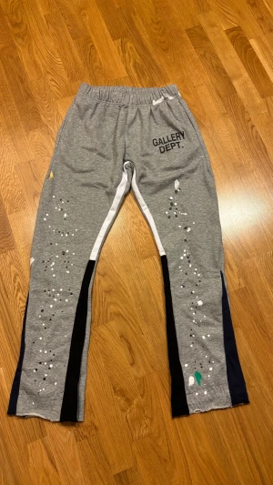 Grå mjukisbyxor från Gallery Dept. - Säljer ett par grå mjukisbyxor från Gallery Dept. med svart tryck på framsidan och färgstänk i vitt, svart och grönt längs benen. Byxorna har elastisk midja, vita och svarta sidopaneler samt råa kanter vid bensluten. Tillverkade i 100% bomull för skön känsla.