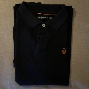 SailRacing pikétröja svart - Svart pikétröja från Sail Racing med orange logga på bröstet och snygga detaljer vid kragen. Klassisk pikémodell med två knappar och mjukt bomullsmaterial som känns skönt mot huden. Perfekt för dig som gillar stilrena och sportiga plagg. Först till kvarn!