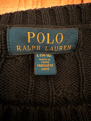 Svart stickad tröja Polo Ralph Lauren - Marinblå stickad tröja från Polo Ralph Lauren med klassiskt kabelstickat mönster och rund hals. Tröjan har långa ärmar och ribbade muddar vid ärmslut och nederkant. På bröstet finns den ikoniska röda Polo-loggan broderad. Perfekt för dig som gillar stilren och tidlös design.        Strorlek: 14-16år.                                                                   Tröjan är marinblå men ljuset på bilder får den att se mörkare ut.