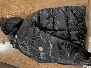 Vinterjacka  - Säljer en svart, glansig pufferjacka från Moncler med huva och quiltad design. Jackan har Moncler-logga på ärmen och coola detaljer som innerfoder med serietidningsmotiv. Perfekt för dig som vill ha en varm och snygg jacka med streetkänsla.