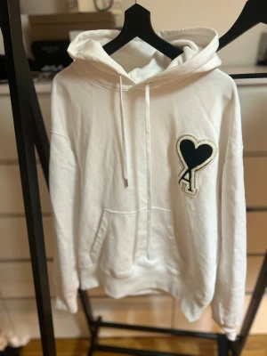 Vit hoodie från Ami med hjärtdetalj - Säljer en vit hoodie från Ami med stor svart hjärtdetalj och bokstaven A på bröstet. Tröjan har huva med snörning, känguruficka och ribbade muddar. Tillverkad i mjuk bomull, perfekt för chill dagar. Ikonisk design som sticker ut.