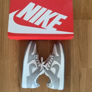 Nike Jordan 1 Low vit/grå sneakers - Säljer ett par Nike Jordan 1 Low sneakers i fräscha vita och ljusgrå toner. Skorna har klassisk snörning, rund tå och platt sula. Ovandelen är i skinn och syntet, med perforerad tåbox för extra andningsförmåga. Perfekta för dig som gillar stilrena och ikoniska sneakers.