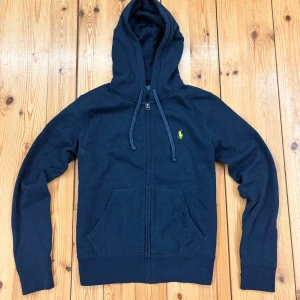 Marinblå hoodie från Polo Ralph Lauren - Snygg marinblå hoodie från Polo Ralph Lauren med dragkedja, huva med snörning och klassisk broderad logga på bröstet. Tröjan har känguruficka och är tillverkad i mjuk bomull, perfekt för chill dagar eller när du vill ha en clean look.