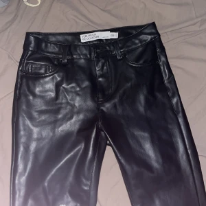 Svarta skinnbyxor från Vero Moda - Snygga svarta byxor i skinnimitation från Vero Moda. Modellen har klassisk femficksdesign, bälteshällor och utsvängda ben. Perfekta för dig som vill ha en edgy look med lite glansig finish. Stängs med dragkedja och knapp framtill.