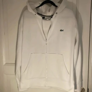 Vit hoodie med dragkedja från Lacoste - Vit hoodie från Lacoste med hel dragkedja och klassisk krokodillogga på bröstet. Tröjan har huva, känguruficka och långa ärmar. Perfekt för en clean och sportig look. Materialet är mjuk bomull som känns skönt mot huden.