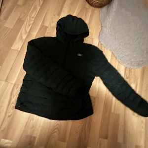 Svart pufferjacka från Lacoste L - Säljer en svart pufferjacka från Lacoste i storlek L. Jackan har huva, dragkedja framtill och det klassiska Lacoste-krokodilmärket på bröstet. Jackan är quiltad och har långa ärmar, perfekt för kyliga dagar. Från mitt perspektiv skulle jag säga att den passar för både folk som har M och S.