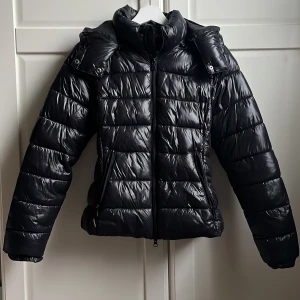 Svart pufferjacka från 157 XS - Svart pufferjacka från 157 i storlek XS. Jackan har glansig yta, huva och dragkedja. Bra skick förutom ett litet hål i ena fickan, men som man inte märker av💗