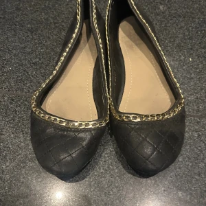 Svarta ballerinaskor med guldkedja - Snygga svarta ballerinaskor med quiltat mönster och guldfärgad kedjedetalj längs kanten. Skorna har rund tå och platt sula, vilket gör dem både stilrena och bekväma. Perfekta för dig som gillar klassiska detaljer med en twist.