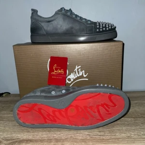 Christian Louboutin grå sneakers med nitar - Grå sneakers från Christian Louboutin i mocka med ikonisk röd sula och coola nitar på tån. Skorna har snörning och en platt sula, perfekt för dig som vill sticka ut med en edgy och lyxig vibe. Snygg och unik detalj med de silvriga nitarna framtill.