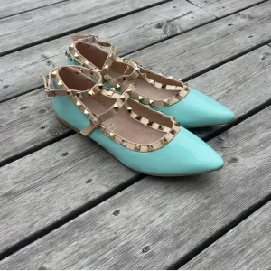 Mintgröna ballerinas med nitar - Snygga ballerina skor i mintgrönt lack med spetsig tå och beigea remmar dekorerade med guldiga nitar. Skorna har en platt sula och stängs med spänne runt vristen. Perfekta för dig som vill sticka ut med en cool detalj och fräsch färg.